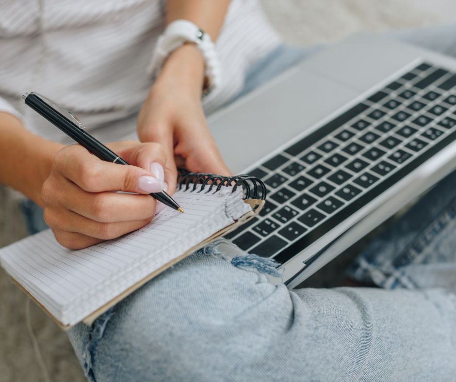 Woman wearing distressed jeans writing in a notebook propped up on her knee with a laptop for the blog post "active voice vs passive voice"