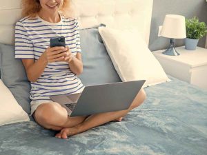 A person sits cross-legged on a bed with a laptop, exploring 2024 content marketing trends on their smartphone while smiling. The bed, draped in a blue blanket, is accompanied by pillows and a lamp on the nightstand.