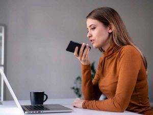 In a cozy corner with her laptop and coffee mug, a woman in an orange top utilizes voice optimization features on her smartphone, illustrating the future of 2024 marketing as she effortlessly navigates through tasks.